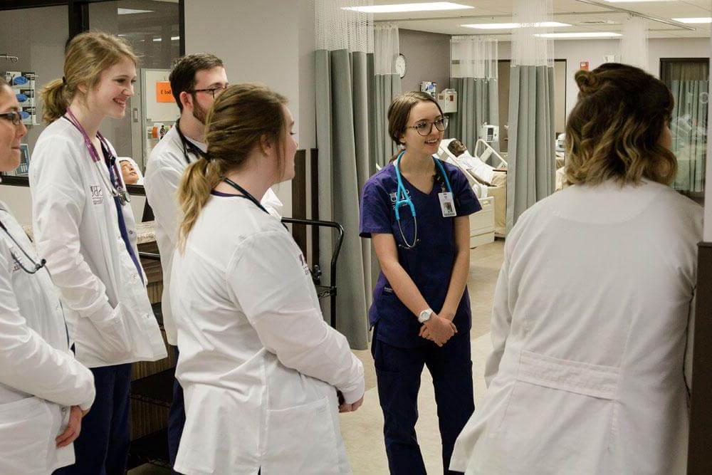 护理专业的学生穿着实习服站在模拟实验室里听教授讲课